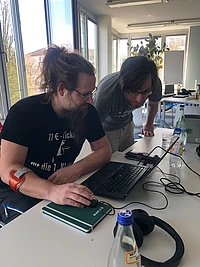 Two people with long hair and glasses look at a laptop on a table in a bright office. There are notebooks, a water bottle, and headphones on the table. One person wears a black shirt and has a red device on their arm.