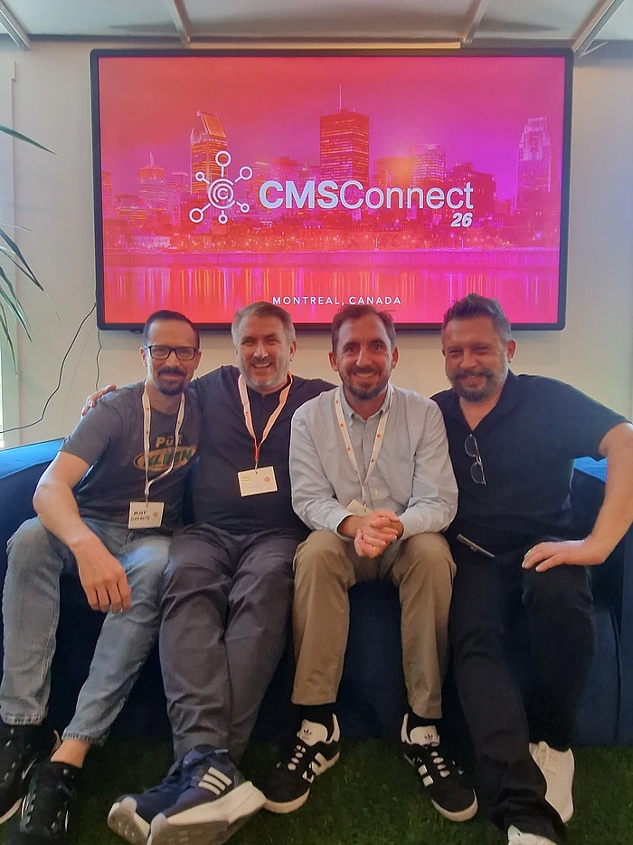 Four men sitting on a couch at CMS Connect 26 conference in Montreal, Canada, with event display on screen behind them.