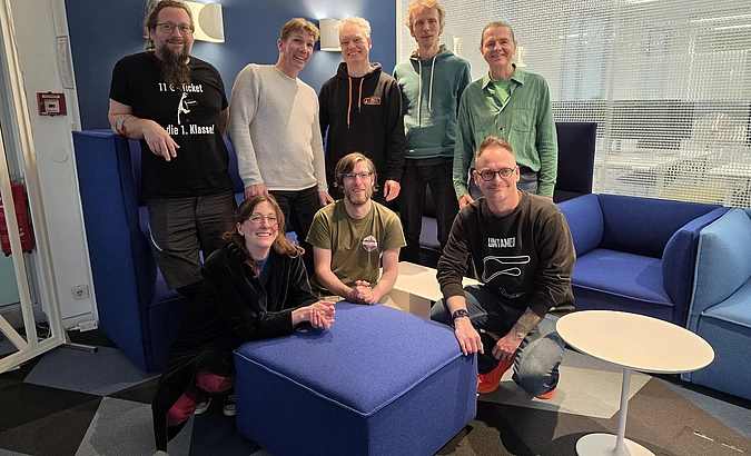 Eight adults pose and smile in a modern lounge area with blue furniture, white tables, and geometric-patterned flooring. Some are sitting on chairs and ottomans, while others stand behind them in casual clothing.