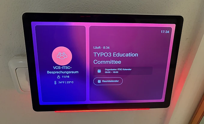 A digital meeting room display shows TYPO3 Education Committee scheduled from 8:00 to 19:00, with the organizer listed as Michael and the current time as 17:34. The screen is bordered by a pink glow.