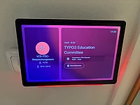 A digital meeting room display shows TYPO3 Education Committee scheduled from 8:00 to 19:00, with the organizer listed as Michael and the current time as 17:34. The screen is bordered by a pink glow.