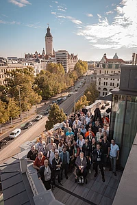 Contao Summit attendees pose for a group photo on a rooftop terrace, with a tree-lined street and Leipzig’s New Town Hall tower behind them.