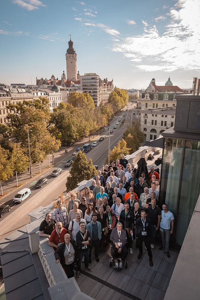 Contao Summit attendees pose for a group photo on a rooftop terrace, with a tree-lined street and Leipzig’s New Town Hall tower behind them.