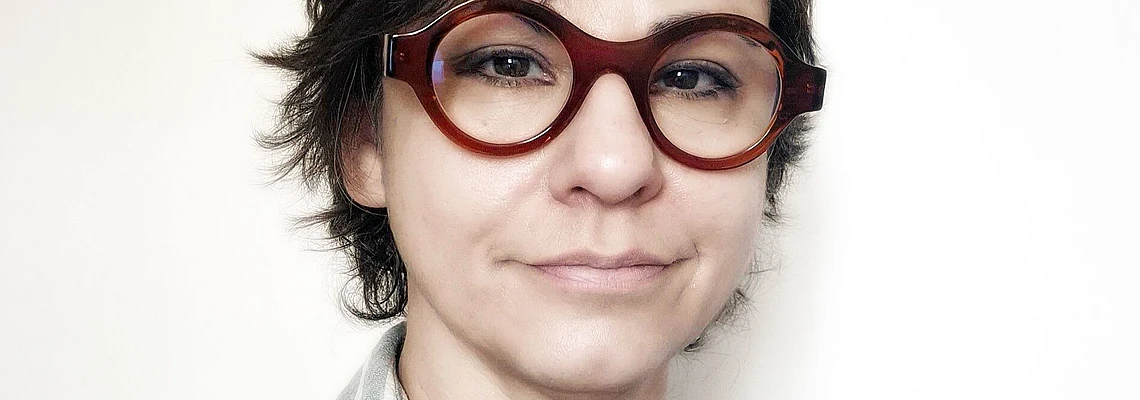A person with short, dark hair and large, round brown glasses is wearing a light gray and white striped shirt and is standing against a plain, light background.