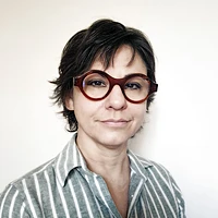 A person with short, dark hair and large, round brown glasses is wearing a light gray and white striped shirt and is standing against a plain, light background.