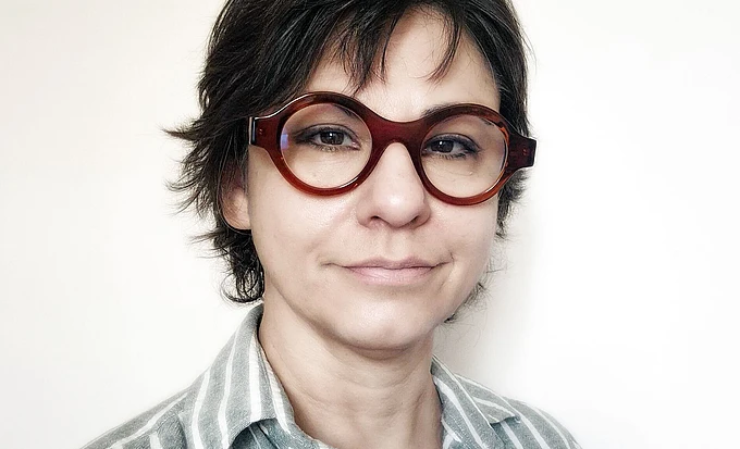 A person with short, dark hair and large, round brown glasses is wearing a light gray and white striped shirt and is standing against a plain, light background.