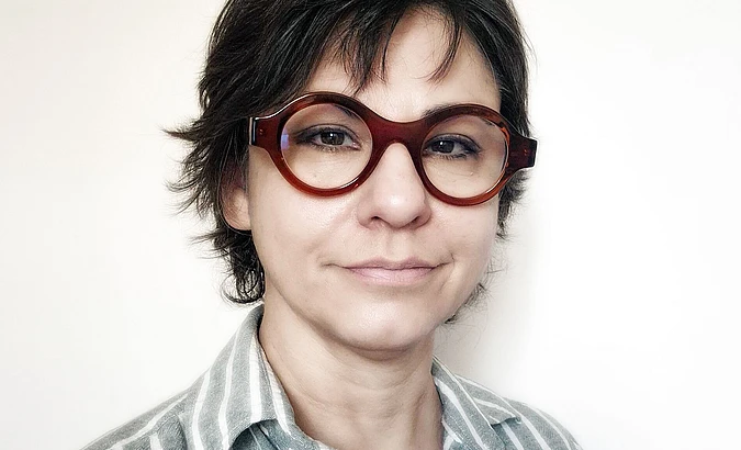 A person with short, dark hair and large, round brown glasses is wearing a light gray and white striped shirt and is standing against a plain, light background.
