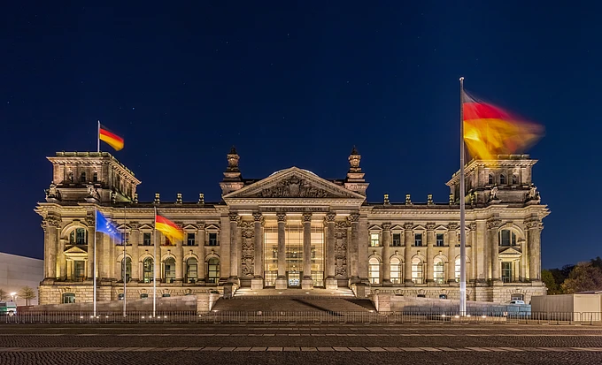 The Reichstag building in Berlin, Germany, is illuminated at night with German and European Union flags flying in front. The neoclassical facade is clearly visible against a dark blue sky.