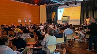 Large audience seated at TYPO3 camp munich in modern auditorium with orange walls watching presentation on yellow screen displaying 'Viel Spaß!!'