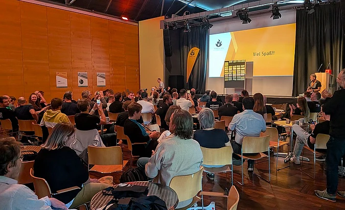 Large audience seated at TYPO3 camp munich in modern auditorium with orange walls watching presentation on yellow screen displaying 'Viel Spaß!!'