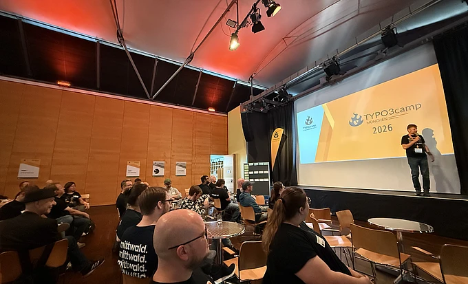 Speaker presenting at TYPO3camp Munich 2026 conference in modern auditorium with audience seated at tables
