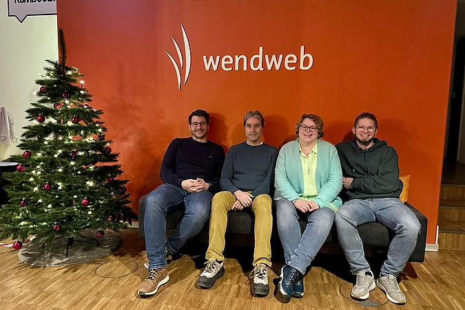 Four people are sitting on a couch in front of a red wall with the wendweb logo. A decorated Christmas tree is on the left side of the image, and all four people are smiling at the camera.