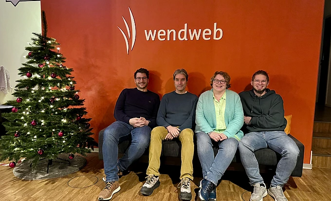 Four people are sitting on a couch in front of a red wall with the wendweb logo. A decorated Christmas tree is on the left side of the image, and all four people are smiling at the camera.