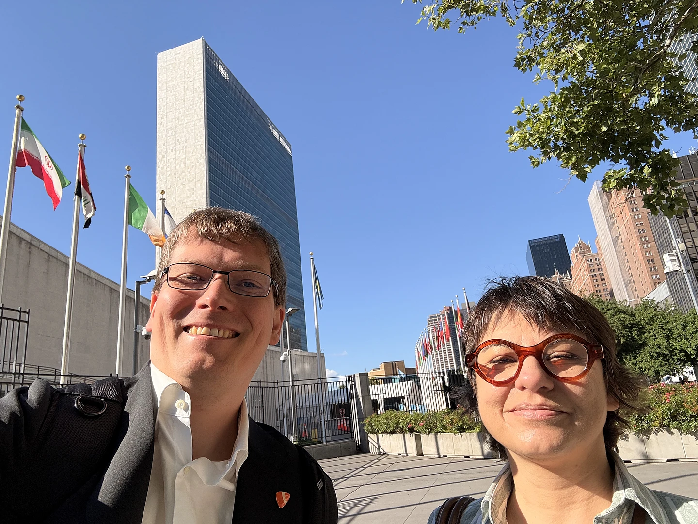 Two people smiling in front of the United Nations headquarters in New York City, with flags from various countries and a tall building visible under a clear blue sky.