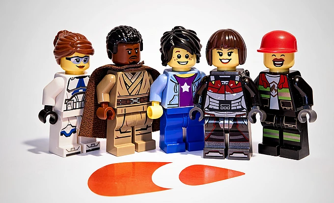 Five LEGO minifigures stand in a row, each with unique outfits and facial expressions, on a white background with an orange TYPO3 logo shape on the floor in front of them.