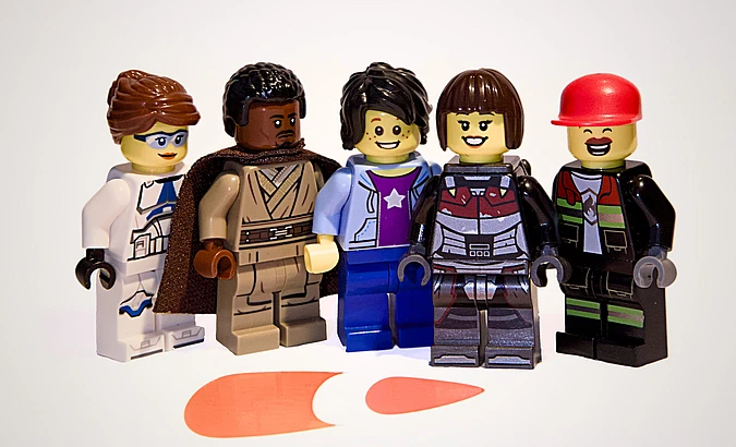 Five LEGO minifigures stand in a row, each with unique outfits and facial expressions, on a white background with an orange TYPO3 logo shape on the floor in front of them.