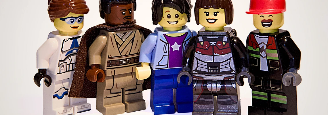 Five LEGO minifigures stand in a row, each with unique outfits and facial expressions, on a white background with an orange TYPO3 logo shape on the floor in front of them.