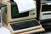 Vintage computer with green monochrome display showing code, keyboard below, and printed documents on top