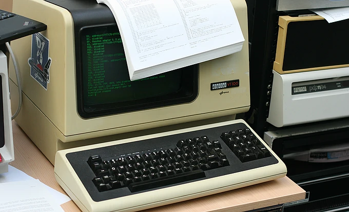 Vintage computer with green monochrome display showing code, keyboard below, and printed documents on top