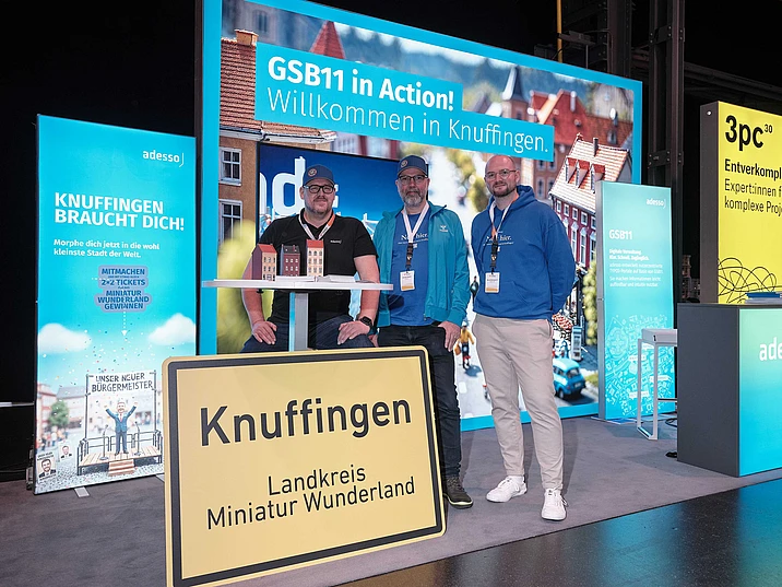 Three men at GSB11 exhibition booth showcasing a fictitious city in Germany called Knuffingen in promotional signage.