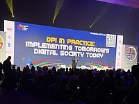 Conference speaker presenting on stage at Global Summit about DPI implementation for digital society, with large audience and blue projection screen