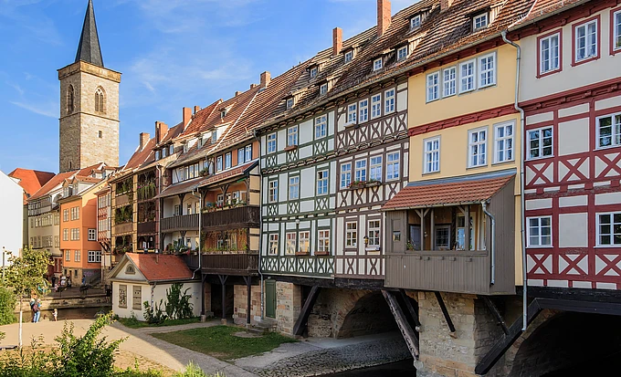 Colorful, timber-framed houses built over a stone bridge span a narrow river, with a church tower in the background under a blue sky. Some balconies feature potted plants and people are walking nearby.