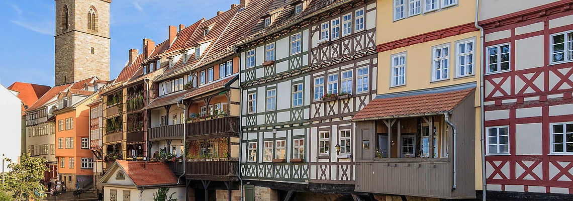 Colorful, timber-framed houses built over a stone bridge span a narrow river, with a church tower in the background under a blue sky. Some balconies feature potted plants and people are walking nearby.
