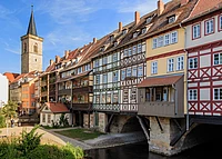 Colorful, timber-framed houses built over a stone bridge span a narrow river, with a church tower in the background under a blue sky. Some balconies feature potted plants and people are walking nearby.
