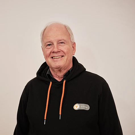 An older man with short white hair smiles at the camera. He is wearing a black hoodie with orange drawstrings and a name badge against a plain, light-colored background.