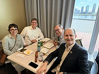 Four people smiling at a dining table with food and drinks, city skyline visible through window behind them