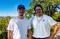 Two men smiling outdoors on a sunny day with green trees and city landscape in background