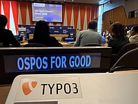 A conference room with people seated; a screen says “UN Open Source Week 2026.” In the foreground, a “TYPO3” sticker is visible, and a sign reads “OSPOS FOR GOOD.”.