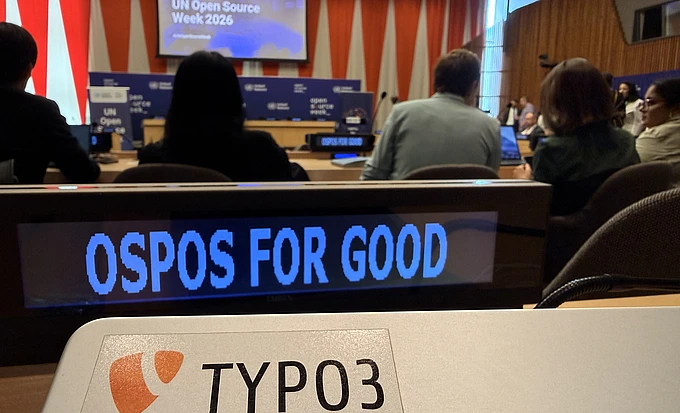 A conference room with people seated; a screen says “UN Open Source Week 2026.” In the foreground, a “TYPO3” sticker is visible, and a sign reads “OSPOS FOR GOOD.”.