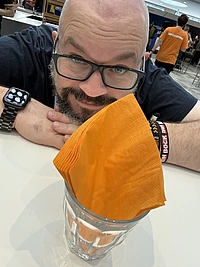Man with glasses and smartwatch ducks behind orange napkins in a glass at indoor event