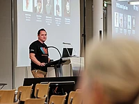 A man stands at a podium giving a presentation in a conference room. He wears a black T-shirt with a colorful design. Behind him, a large screen displays profile pictures and names. Some empty chairs are visible in the foreground.