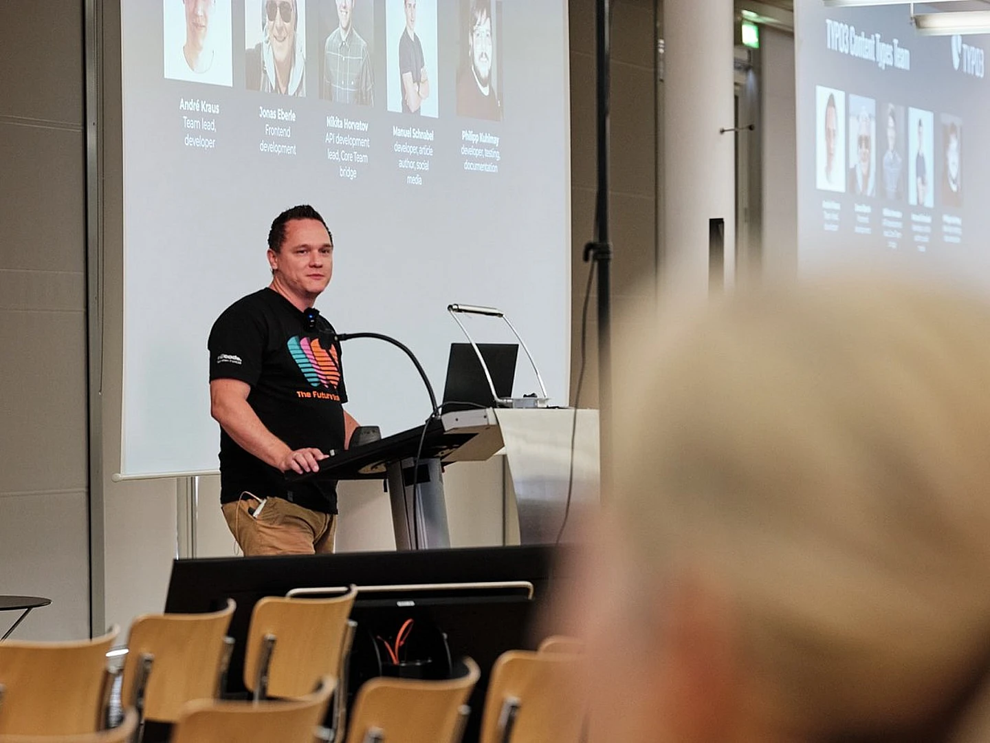 A man stands at a podium giving a presentation in a conference room. He wears a black T-shirt with a colorful design. Behind him, a large screen displays profile pictures and names. Some empty chairs are visible in the foreground.