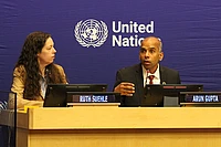Two people sit at a conference table with United Nations signage behind them. The man on the right is speaking and gesturing, while the woman on the left listens. Nameplates read “Ruth Suehle” and “Arun Gupta.”.