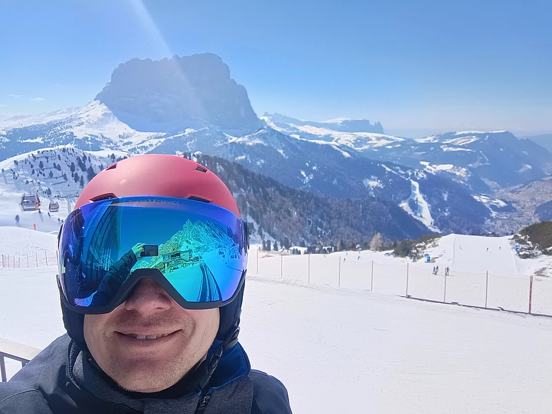Person wearing a red helmet and reflective ski goggles smiling on a snowy mountain slope with ski tracks and distant peaks visible under a clear blue sky.