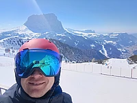 Person wearing a red helmet and reflective ski goggles smiling on a snowy mountain slope with ski tracks and distant peaks visible under a clear blue sky.