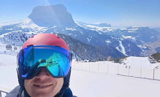 Person wearing a red helmet and reflective ski goggles smiling on a snowy mountain slope with ski tracks and distant peaks visible under a clear blue sky.