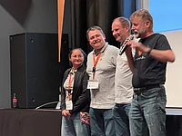 Four people on stage at a conference, wearing ID badges, one holding a microphone during a presentation