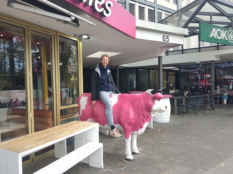 A person sits on a life-sized pink and white cow statue outside a shop with glass doors. People are seated at tables nearby. The scene is urban, with store signs and a street visible.