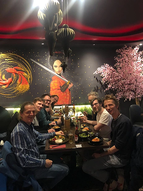 Seven people dine at a restaurant with Asian decor, including a mural of a woman with a sword and a cherry blossom tree. The group is smiling around a table with food and drinks.