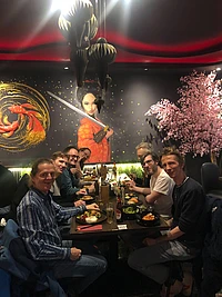 Seven people dine at a restaurant with Asian decor, including a mural of a woman with a sword and a cherry blossom tree. The group is smiling around a table with food and drinks.