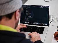 A person photographed close from behind is typing some code into their laptop. code sprint community developer laptop typo3 code