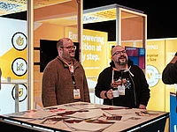 Two men with conference badges stand smiling at a TYPO3 exhibition booth with bright yellow displays, flyers on the table, and one man holding a white cup. The background features text and tech-themed graphics.
