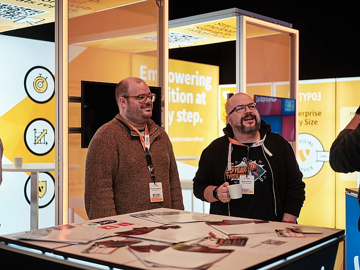 Two men with conference badges stand smiling at a TYPO3 exhibition booth with bright yellow displays, flyers on the table, and one man holding a white cup. The background features text and tech-themed graphics.