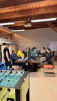 Several people sit at computer desks working on desktop monitors in a modern office with exposed wooden beams. Two others stand nearby. A foosball table is in the foreground and shelves and a whiteboard are in the background.