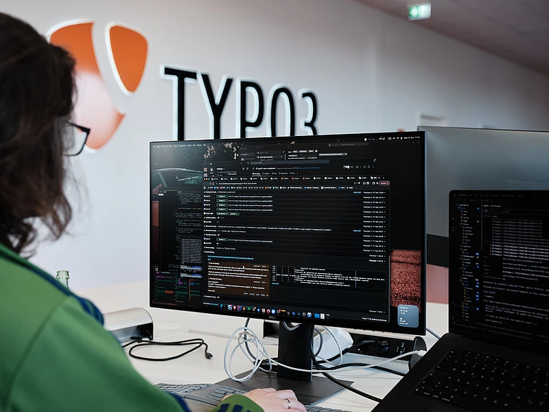 A person is working at a computer with code and terminal windows open on the screen, sitting in a modern office space with a large TYPO3 logo on the wall in the background.