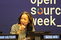 A woman speaks into a microphone at a conference table, with a blue open source week banner behind her and nameplate reading SACHIKO MUTO in front.